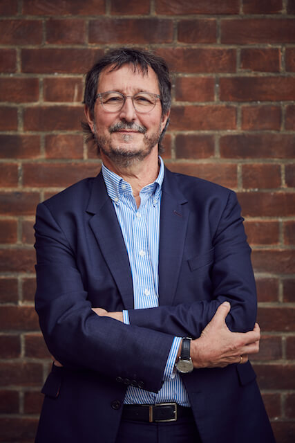 Professor John Last OBE profile picture smiling with brick wall behind