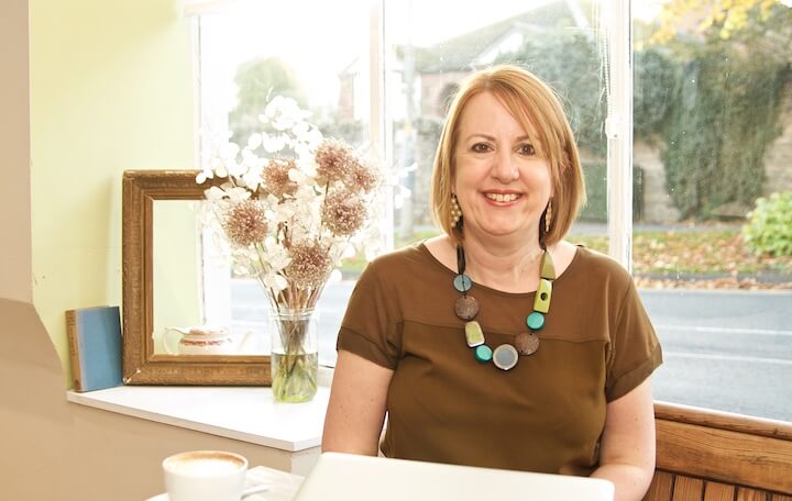 Lucy Eckley smiling profile picture sitting with coffee in front of window flowers mirror